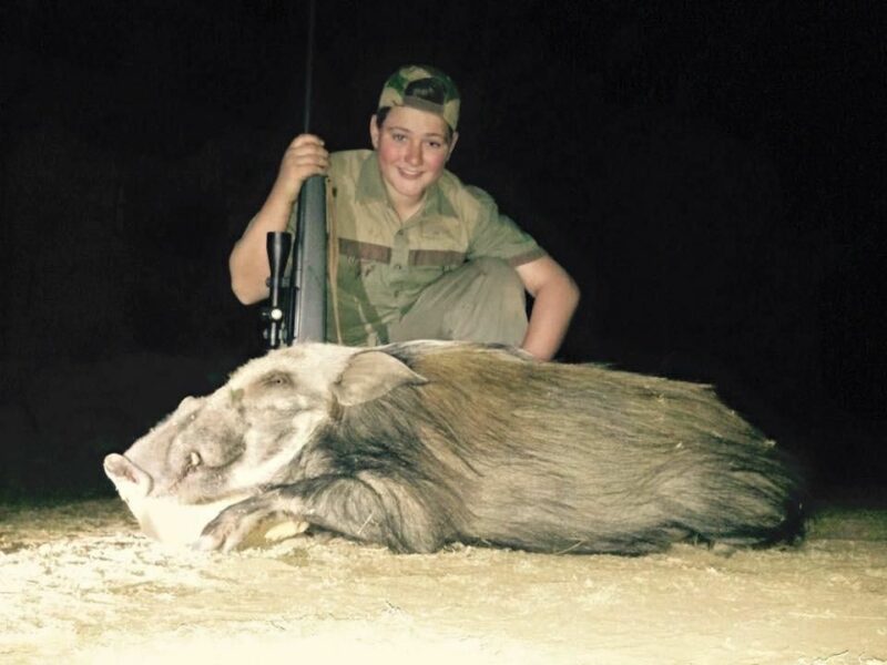 Boy posing with a rifle, showing the bushpig he shot.